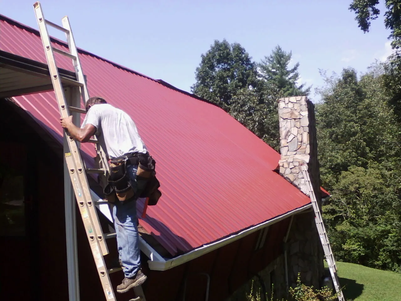 Roofer on a ladder beside a red metal roof and stone chimney — Soffit & Fascia Repair by Roofing Brothers in Waucoma